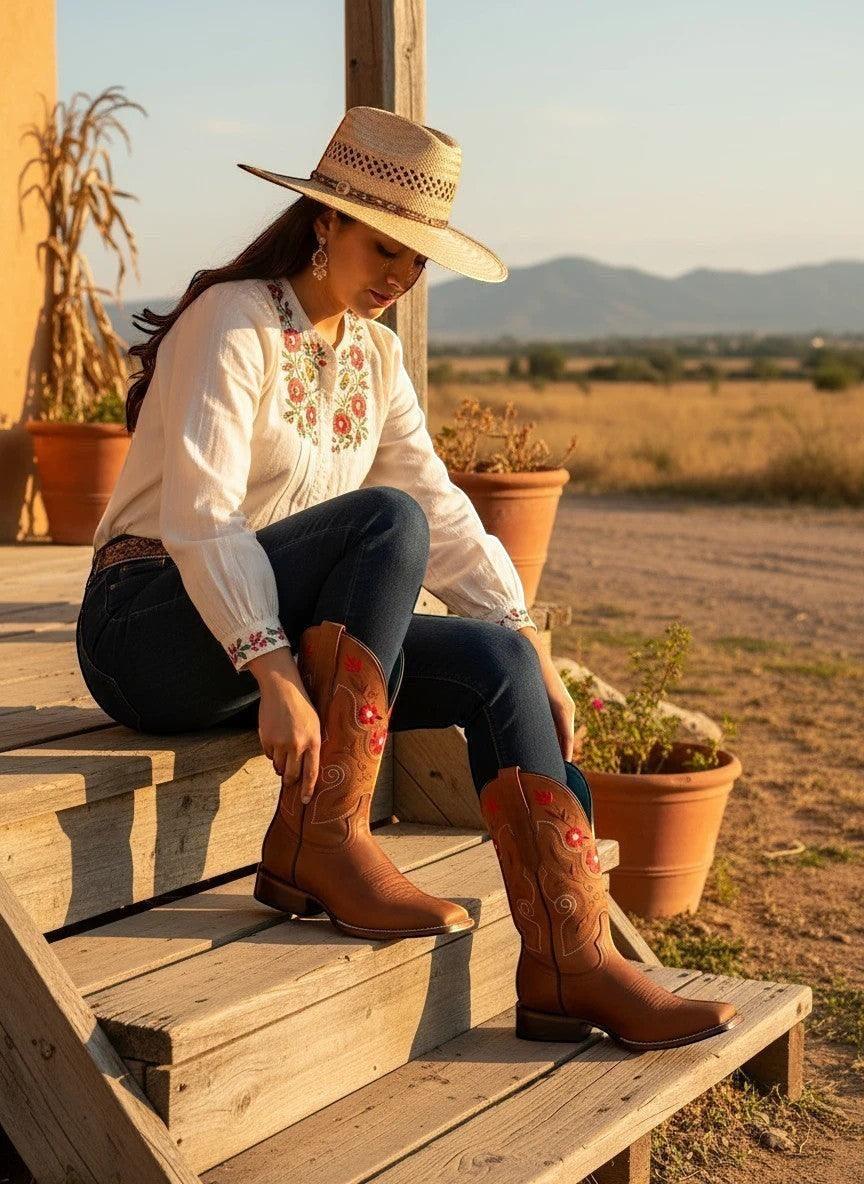 Mujer con botas vaqueras RAVAC de piel bovino color shedron y sombrero, sentada en un porche rústico al atardecer.